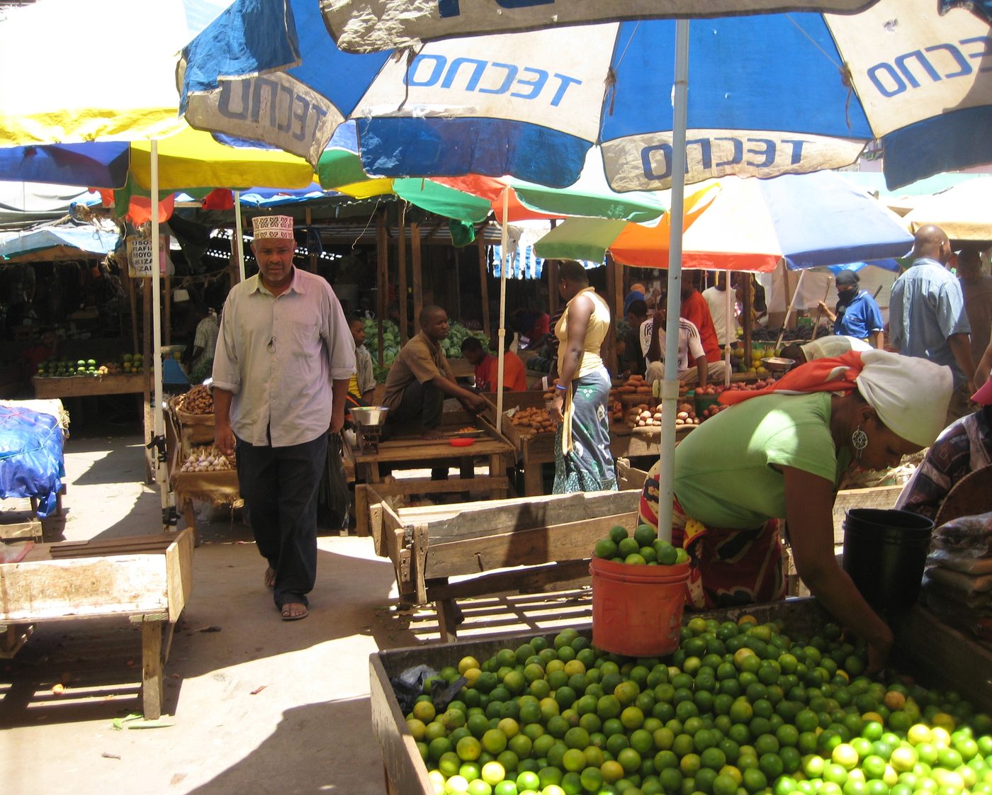 A busy covered market lane under coloured umbrellas, with a man walking between wooden stalls and a woman arranging a large pile of limes in the foreground.